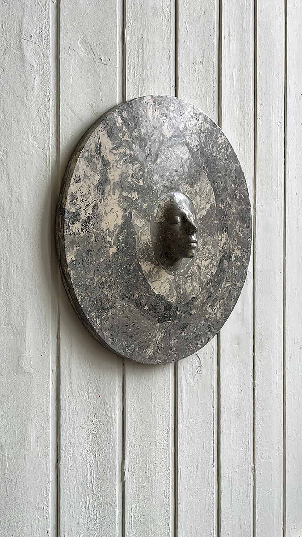 Round grey marbled plaster relief with a cast human face emerging from its centre, shown installed on a white wall in bright light.