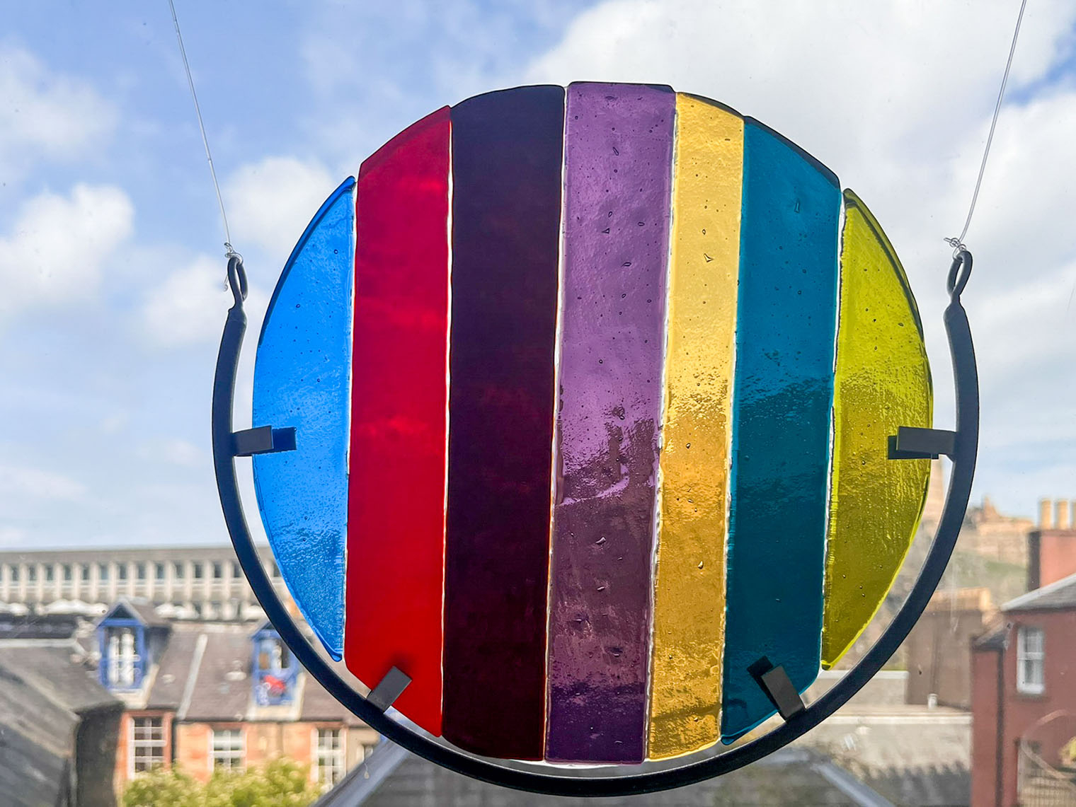 Circular fused glass artwork in a metal frame, hanging in a window and catching natural light, with the outdoor landscape visible behind it.