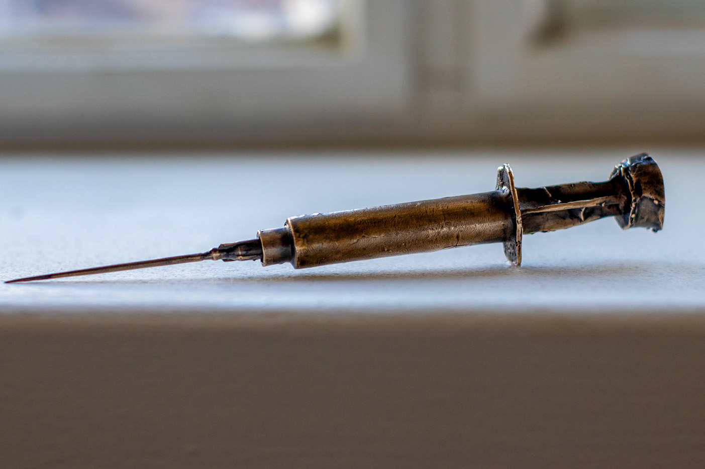 Bronze cast of a syringe, 17 cm long and 1 cm wide, placed on a white window shelf.