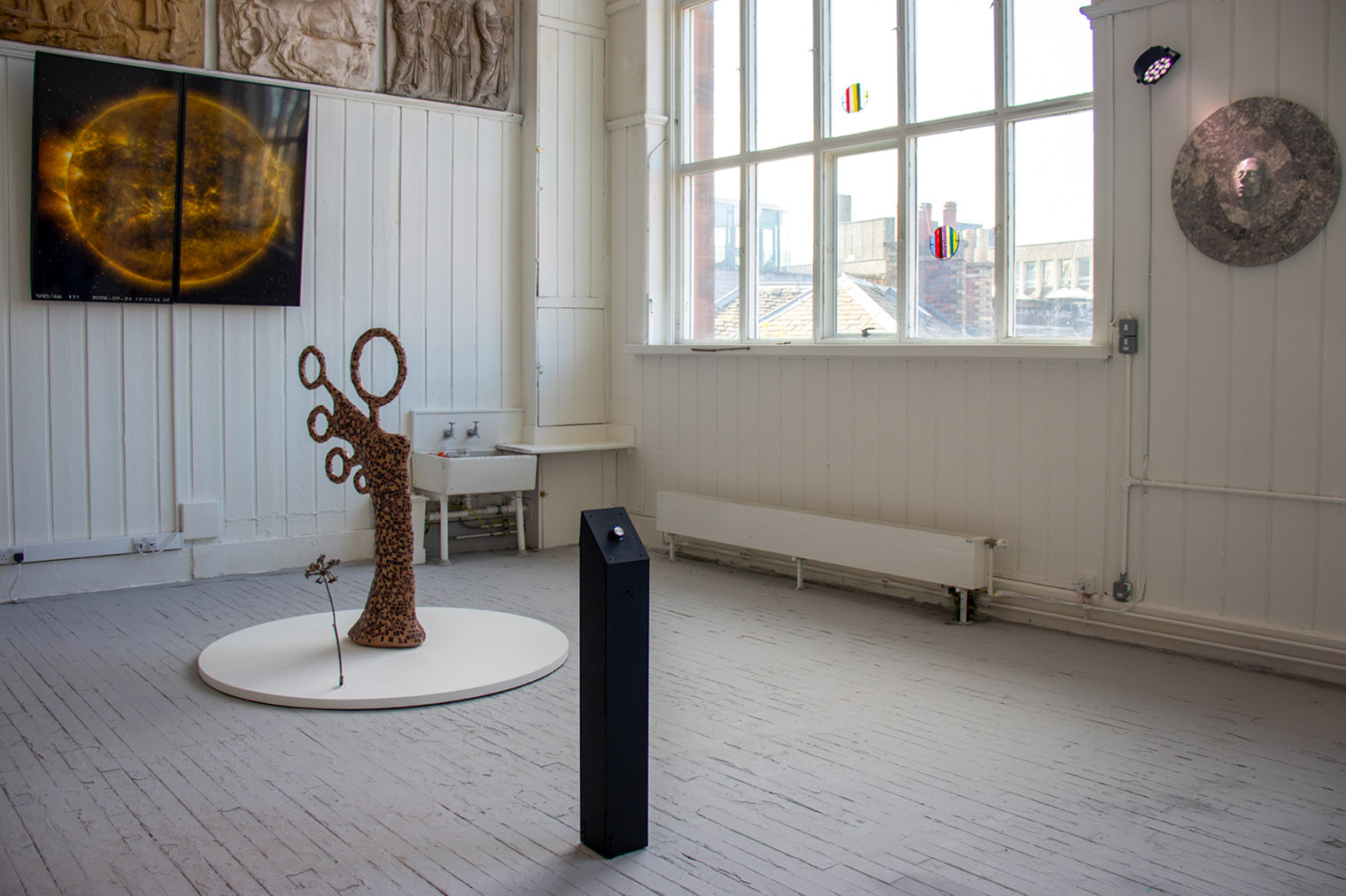 Installation view of an abstract mosaic figure in brown ceramic tiles on a white round plinth, beside a bronze cast of hogweed. Screens display a sun image, and windows feature two circular pieces of fused glass.