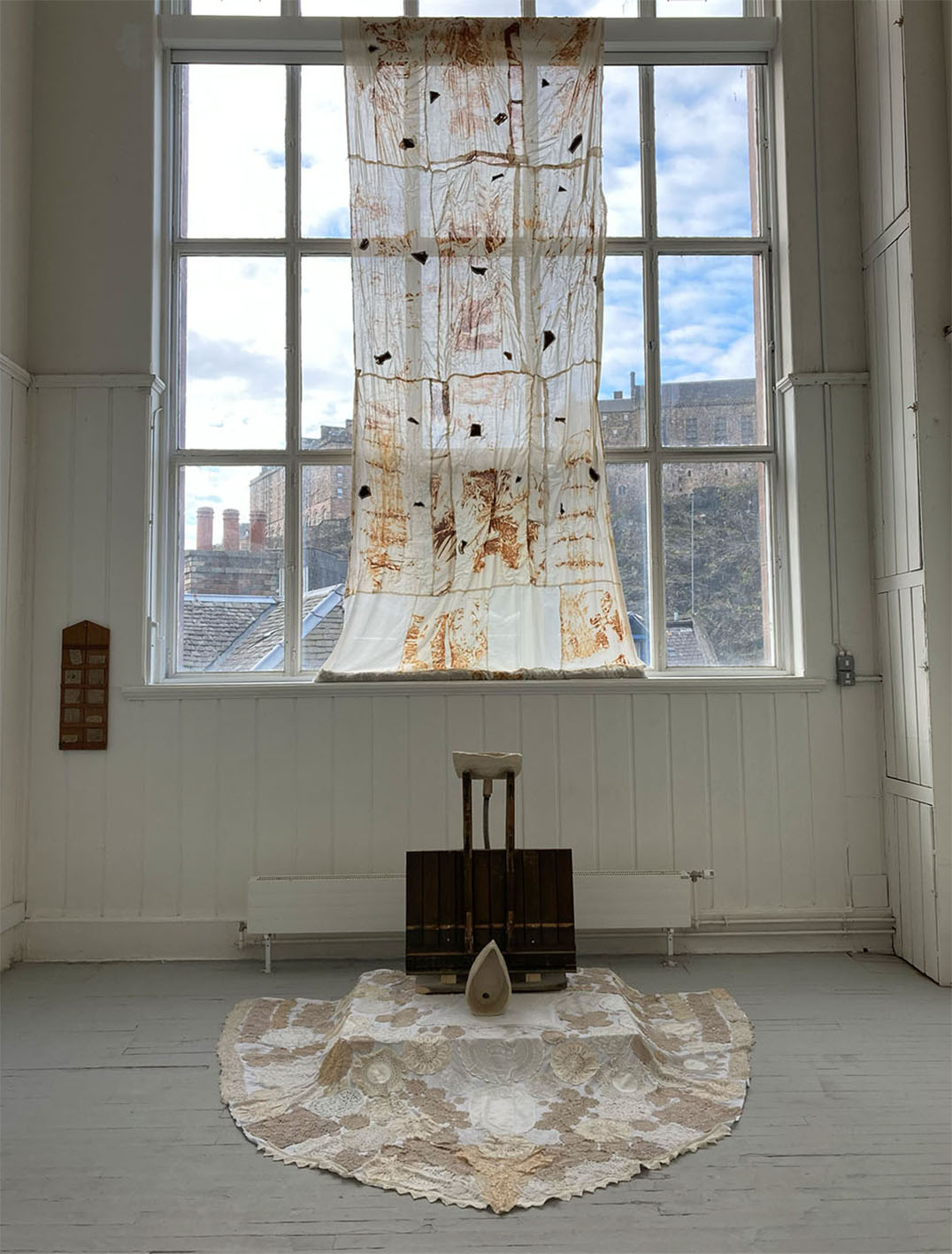 Image of sculptural instatllation including textiles hanging from a window, and a rug and wooden structure on the floor.
