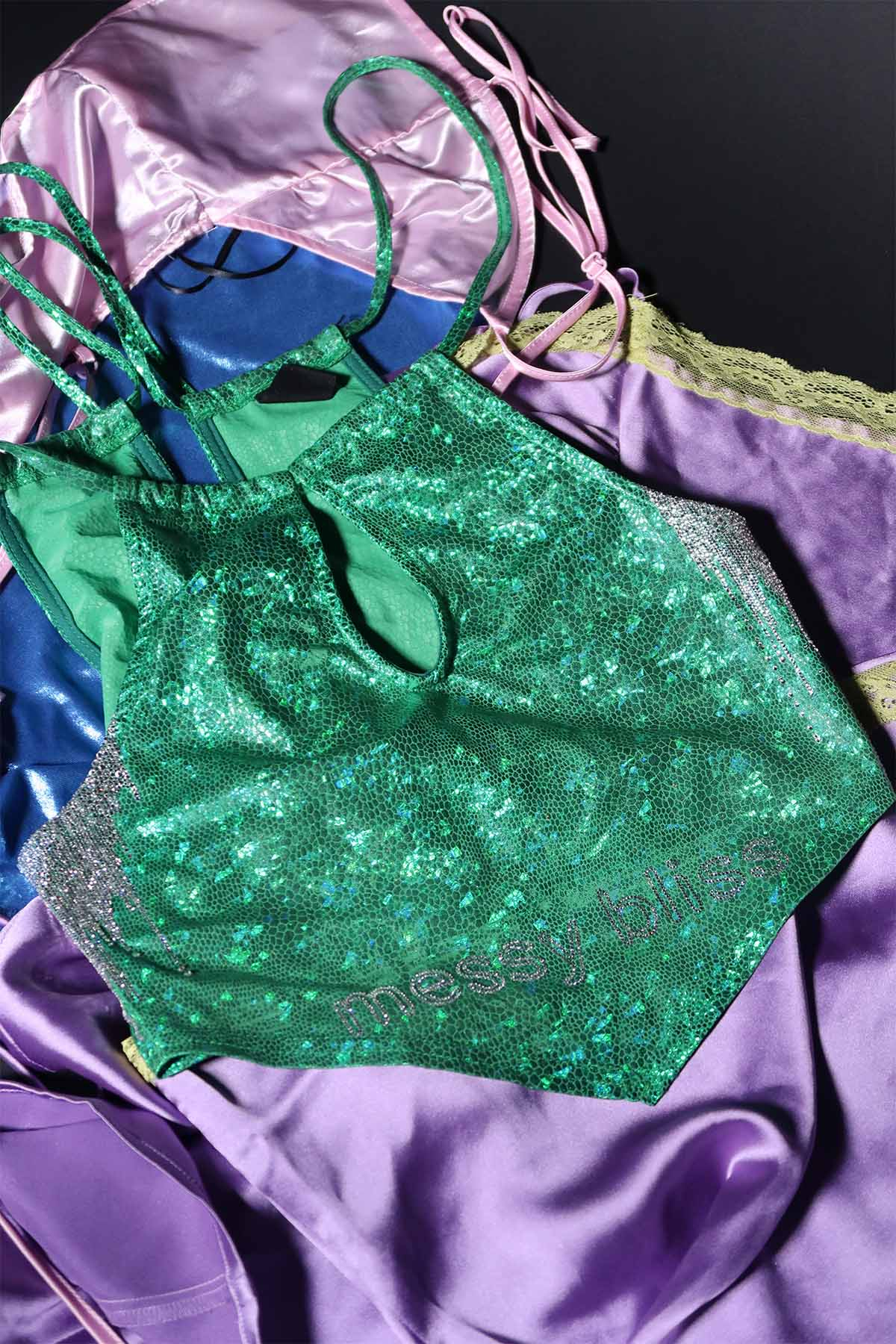 A metallic green halter-neck top is at the top of a pile of clothing on a black background. It has a slogan in silver rhinestones that says ‘messy bliss’. The underarm areas have lines of silver rhinestones. The rhinestones are saturated at the top of the underarm, but become individual droplets as they move down the side of the top.