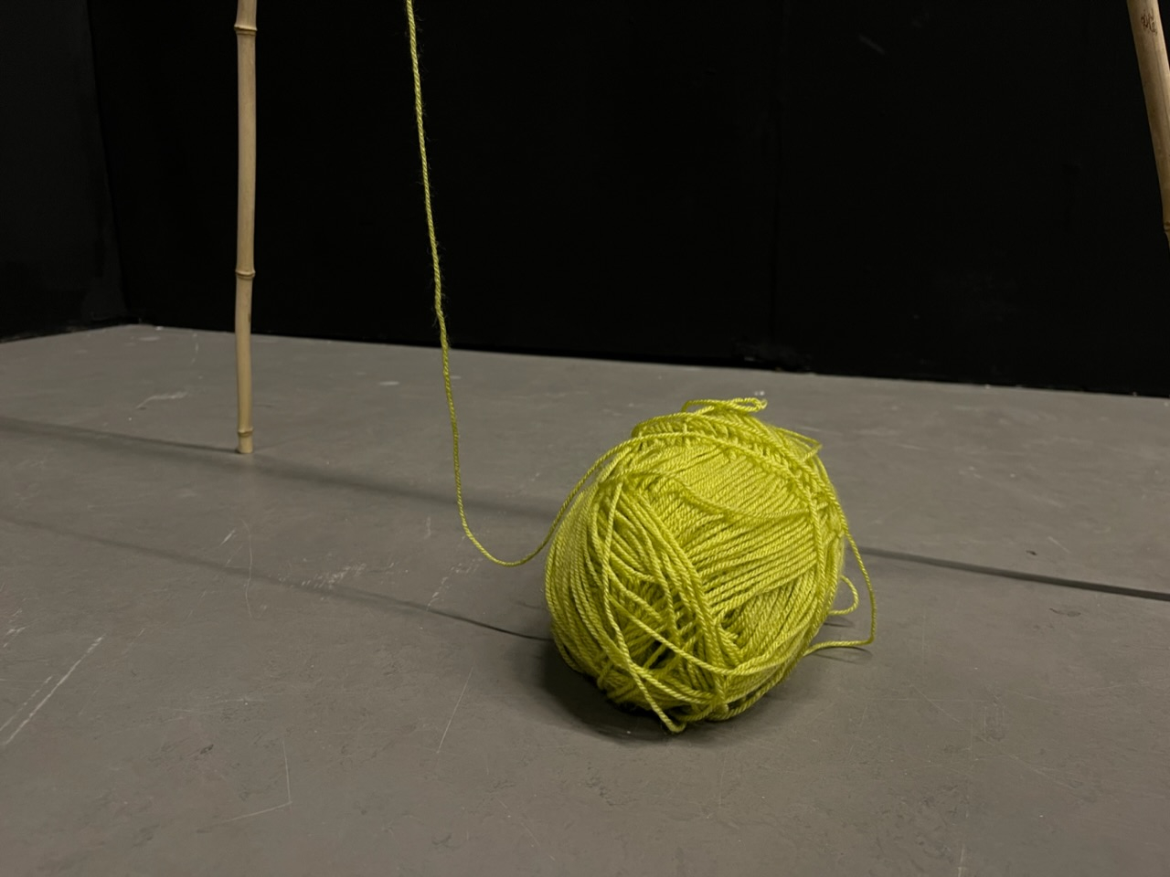 Ball of green acrylic yarn and upright bamboo stick on grey floor, leading up off the top of the image