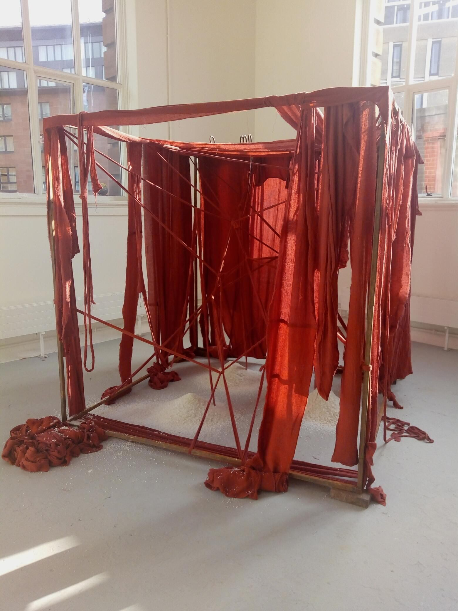 A 2 metre by 2 metre rusted steel frame with internal structure of red fabric criss-crossed and draped and wound with red fabric bandages. There are 3 piles of salt within the structure and 4 upstanding metal hooks at the rear and to the left.