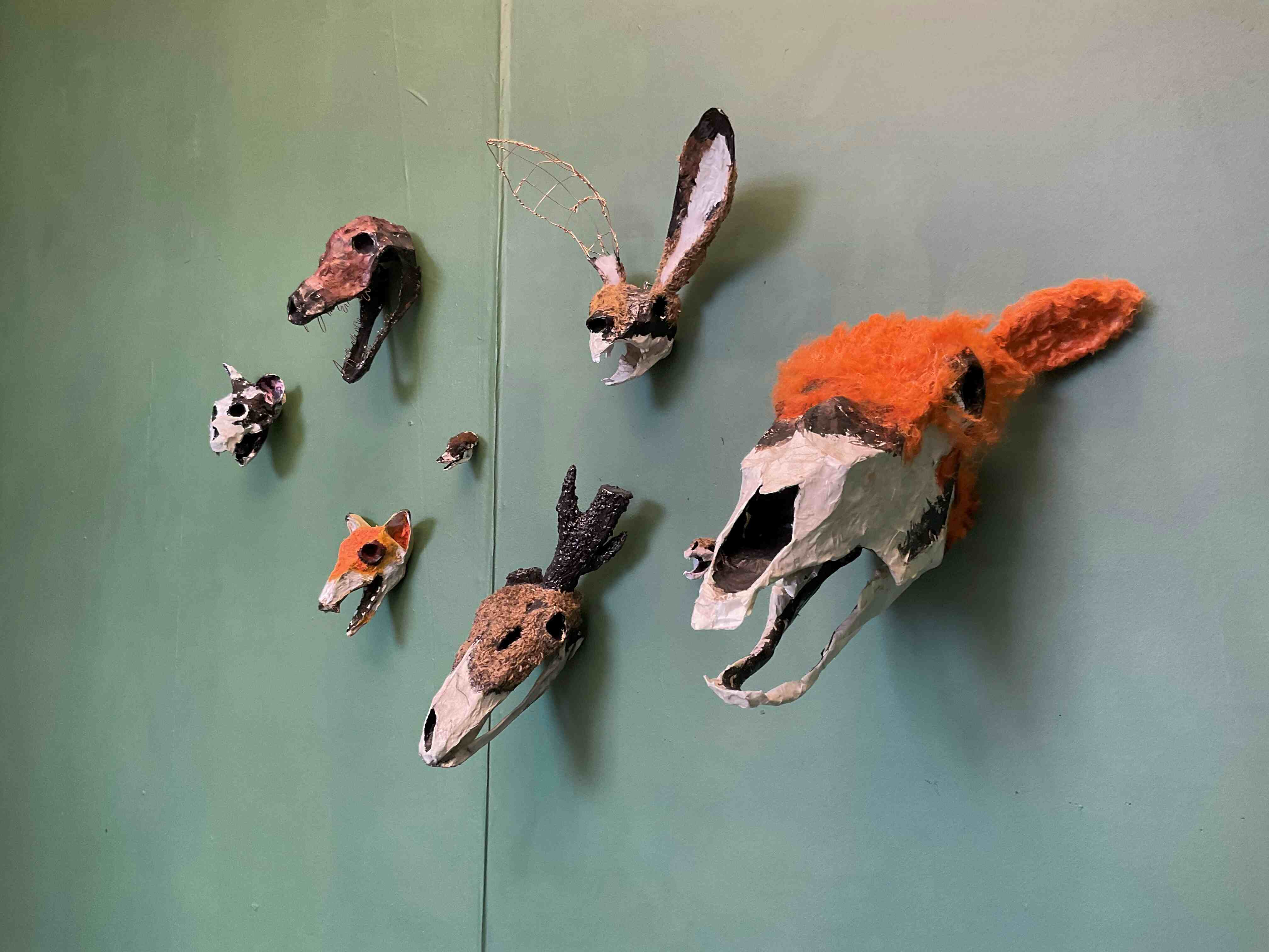 Animal head sculptures on a sap green background. The animals (a highland cow, hare, dog, cat, fox, shrew, deer and mouse) are all in decayed states showing parts of their skulls but retain some wool 'fur'. 