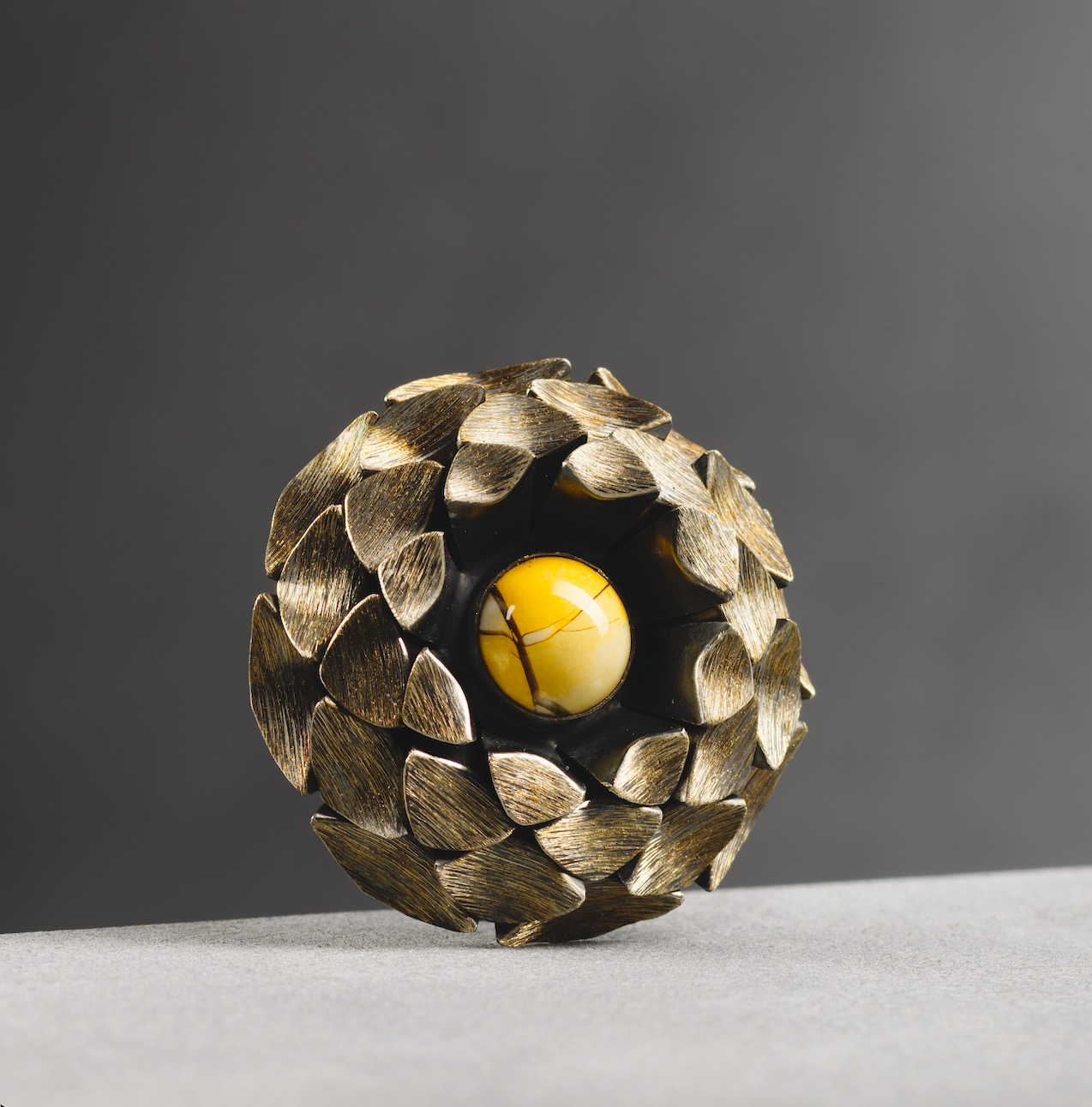 Circular layered brooch made of folded silver metal elements with gold arranged in a pinecone-like structure, centred with a polished brown jasper stone and featuring overlapping geometric forms inspired by natural growth patterns.
