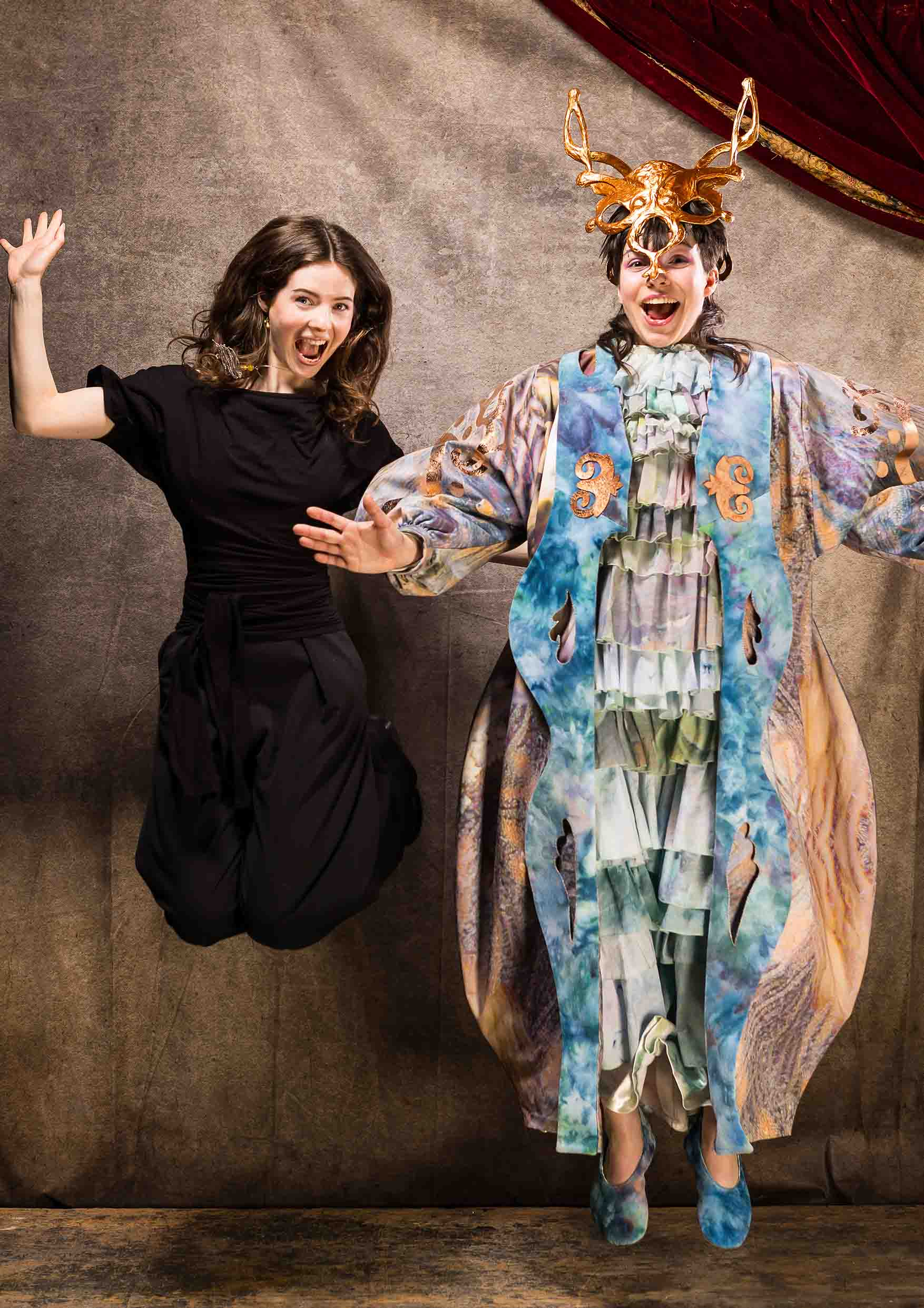Two people jumping, one in all black and one wearing long colourful coat and metallic headpiece