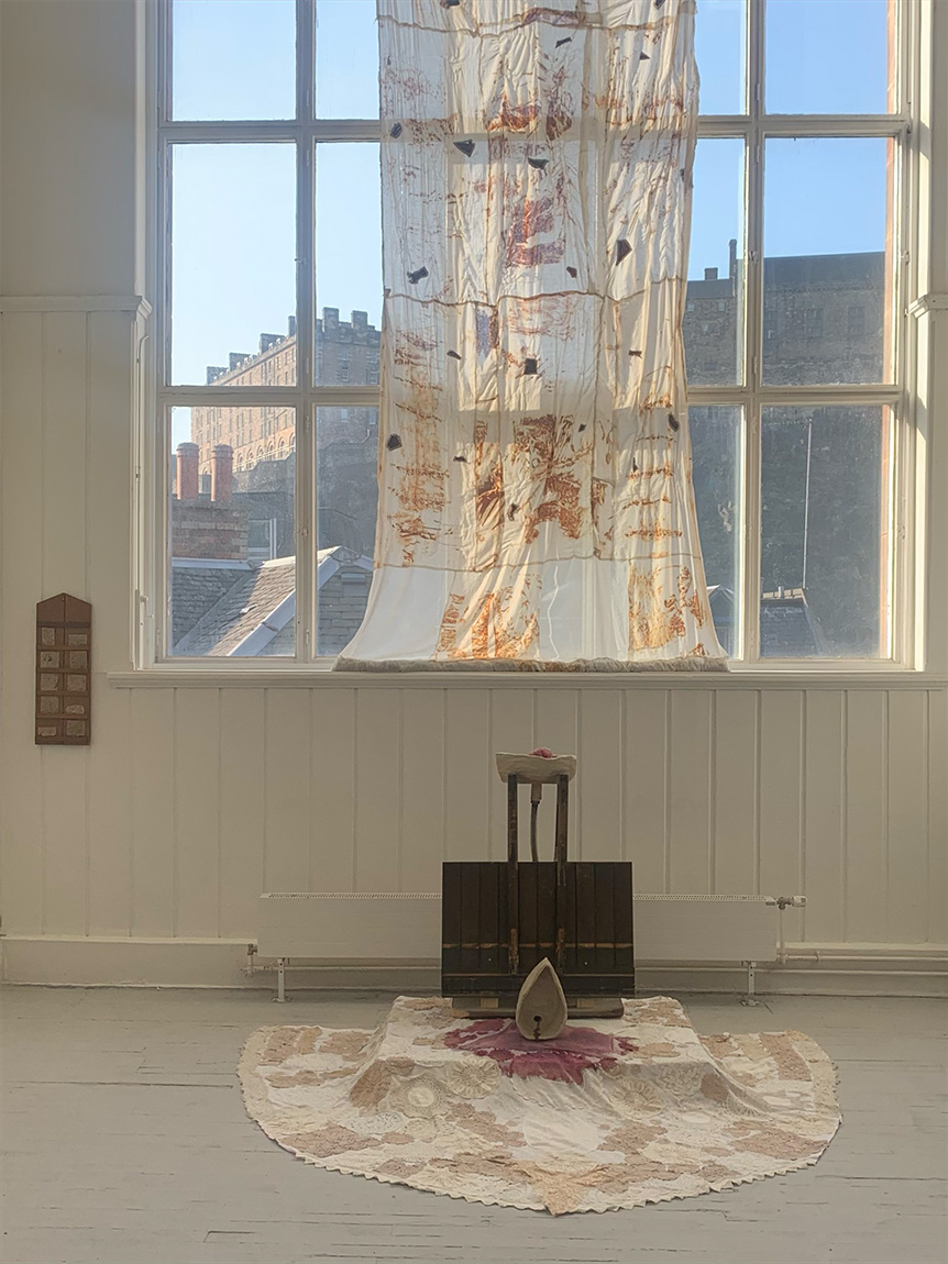 image of sculptural instatllation including textiles hanging from a window, and a rug and wooden structure on the floor.