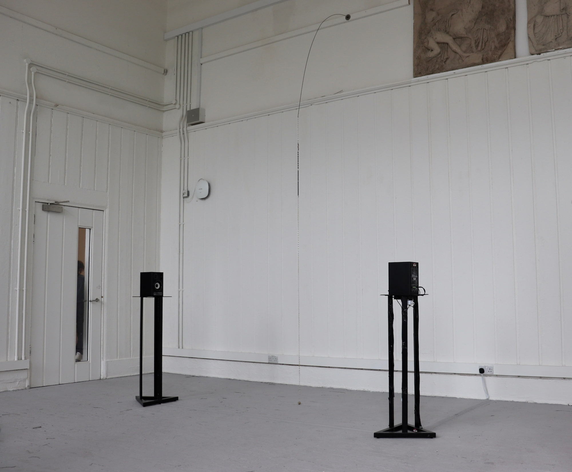 two speakers on stands facing each other, a meter a part. a beaded sculpture hanging from an iron rod from the wall.