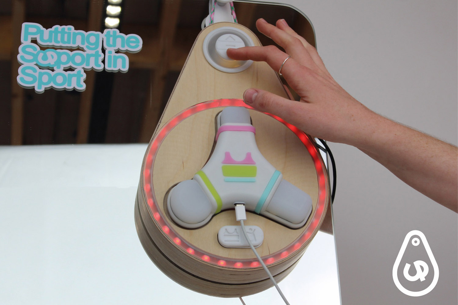 A hand reaches out to touch a button on an oval shaped object which is made of wood. There is a 3-pronged device in the centre which sits in the centre of a circle of red LED lights. The text reads 'putting the support in sport'.